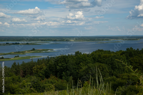 A view of the Dnipro river, with dense green forest on the near shore in the foreground, islands on the water, a distant shore beyond the river, and blue sky with fluffy white clouds above.