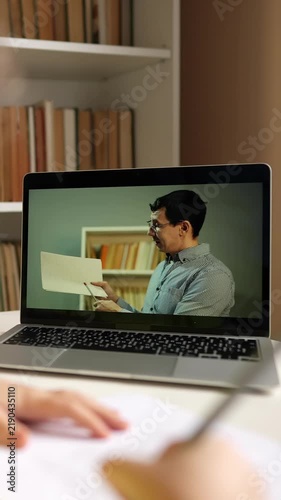 A man is speaking on a laptop during a virtual class. A student can be seen taking notes while the man presents information in a home setting.