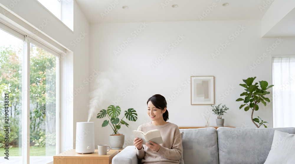 Fototapeta premium Relaxed woman reading book on sofa near ultrasonic aroma humidifier releasing hydrating mist in living room