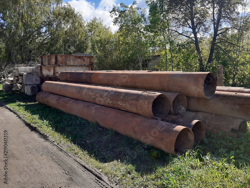 Stack of Rusty Metal Pipes on Grass Outdoors. Used cylindrical pipes are laid out at a construction site next to concrete blocks. Corroded, dirty, elongated iron structures are piled up near the trees