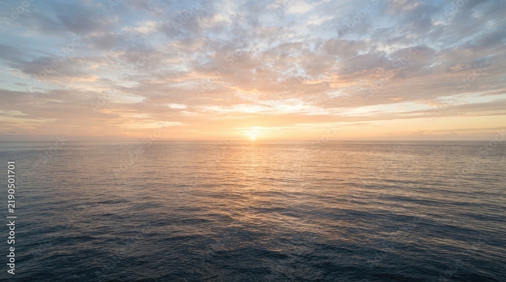 Obraz premium The sun rises over the ocean, casting a warm glow on the water and sky. Clouds are scattered across the horizon, reflecting the colors of the sunrise.
