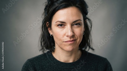 A woman with dark hair and a serious expression looks directly at the camera. She is wearing a gray sweater.