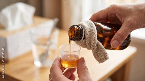 Hands pouring medicine into a glass measuring cup from a Cough syrup bottle with scarf