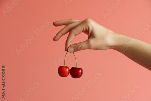 Female Hand Holding Two Fresh Red Cherries by Stems Against Pink Background