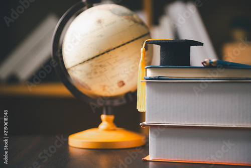 Fotografie Graduation cap placed on stacked books beside a large classroom globe, representing education, geography, global learning, academic achievement, knowledge, study, scholarship, and school success