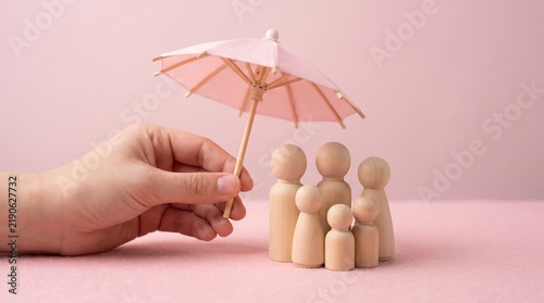 Family life insurance concept, wooden peg figures under pink umbrella, hand protecting family with insurance coverage