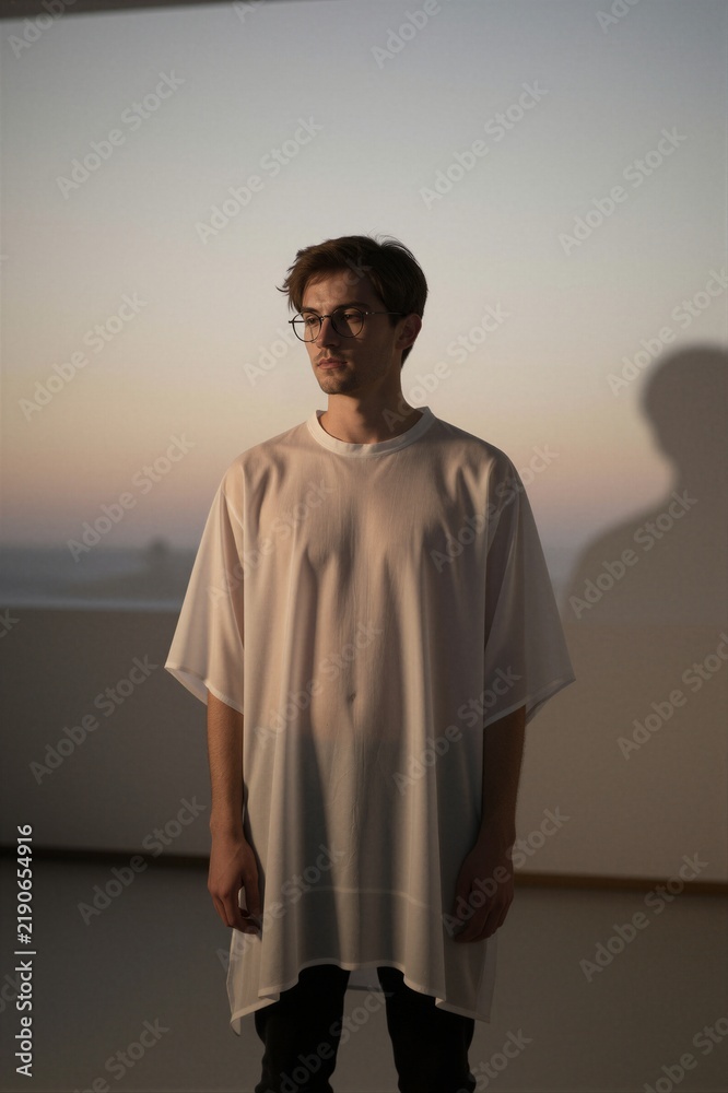 Obraz premium A young man stands in a serene indoor space, wearing an oversized white T-shirt and black pants, gazing out at a warm gradient sky through round glasses.