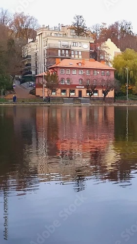 Vertical panoramic shot from city lake waterfront of calm mirror water surface and European style residential buildings among colorful autumnal foliage trees at autumn day. Scenic fall season scenery.