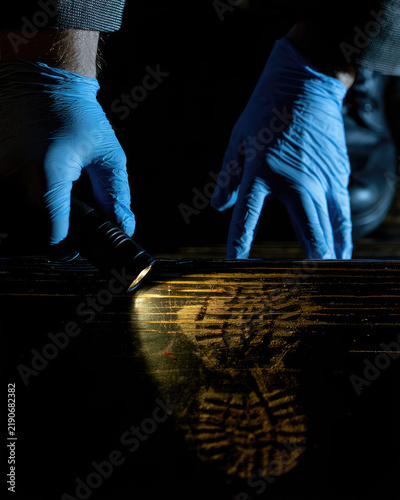 Forensic investigator in blue gloves examining faint bloody shoeprint on dark wooden stair with angled flashlight, revealing tread detail under high contrast crime scene lighting