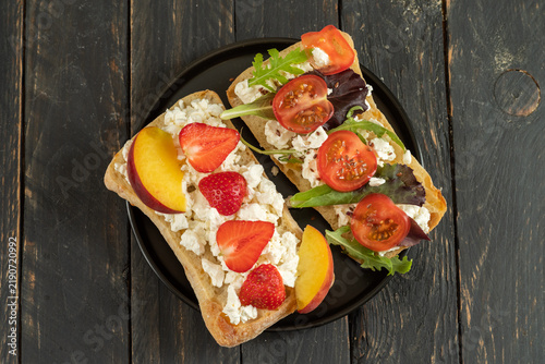 Two open sandwiches on a black plate: one with tomatoes and arugula, the other with cottage cheese, peach, and strawberry. Served.