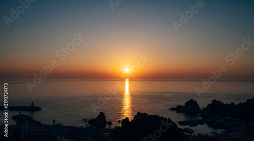Beautiful golden sunset over a calm ocean horizon with dark rocky coastline and shimmering water reflections