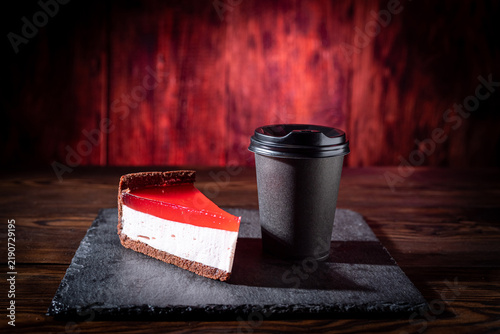 A slice of cheesecake topped with red berry jam alongside coffee in a black craft paper cup on a rich red background. Delicious dessert and coffee.