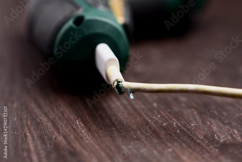Damaged frayed power cord of an electric tool on a wooden surface. Broken white charger cable insulation with exposed wires.