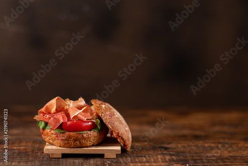 A gourmet sandwich made with a whole grain seeded bun, jamon, ripe cherry tomatoes, and fresh greens on a rustic wooden background.