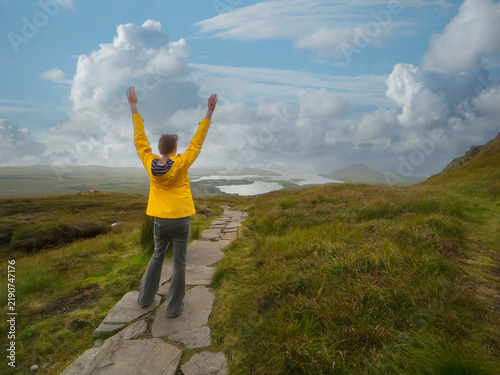 Bild auf Leinwand Person in a bright yellow raincoat stands on a stone path, facing a vast, rugged landscape