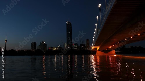 The glowing bridge and modern Vienna skyline reflect gracefully on the dark Danube river at night. Vibrant urban landscapes inspire dynamic economic growth and celebrate advanced city infrastructure.