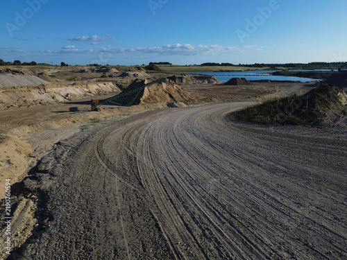 Aerial industrial gravel pit with sand piles, conveyor system, curved haul road and water area in open extraction landscape