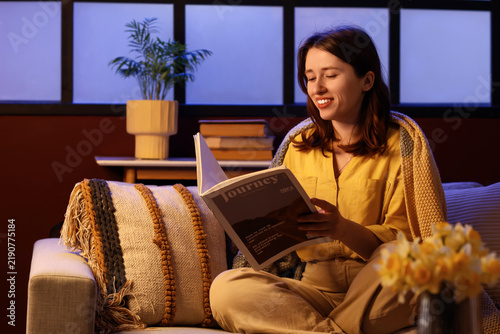 Young happy woman with soft plaid reading magazine on sofa near window in eve...