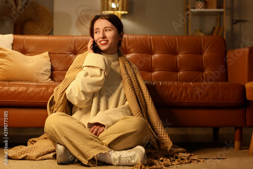 Young happy woman with soft plaid talking by mobile phone on floor in evening...