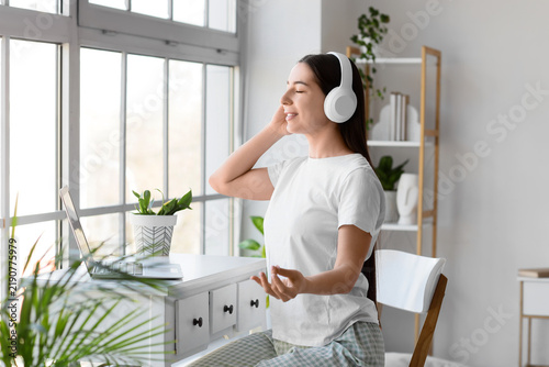 Beautiful young woman with headphones and laptop meditating at table in bedroom