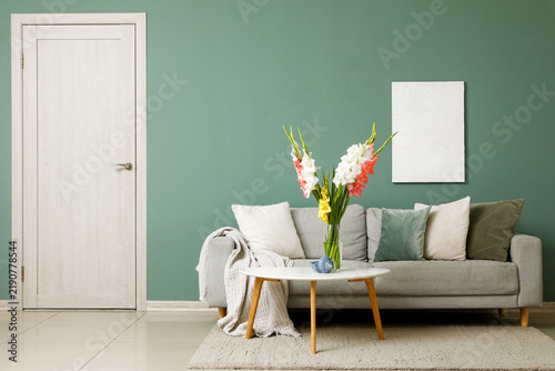 Comfortable sofa, table and vase with beautiful gladiolus flowers in interior...