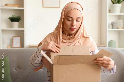 Young Muslim woman opening parcel box on sofa at home