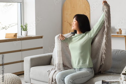 Young Asian woman with grey plaid stretching on sofa at home