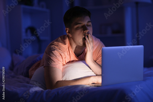Handsome young man with laptop yawning while lying in bed at night