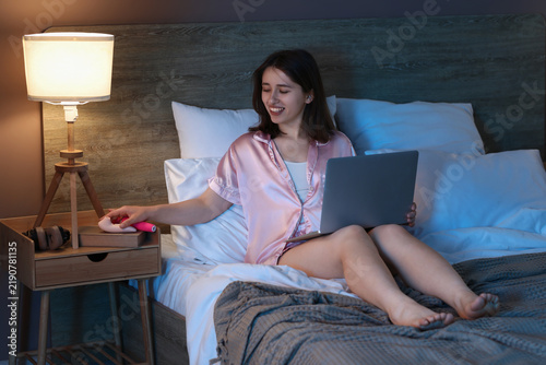 Young pretty woman with laptop taking sex toys from bedside table in bedroom ...