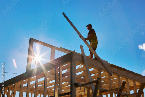 Принти на полотні Installing roof rafters, young builder works on timber frame house while assembling wooden roof structure during construction