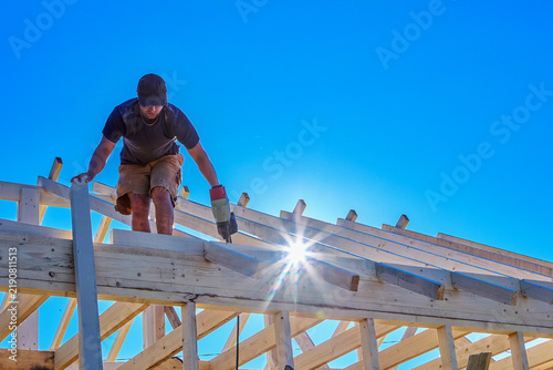 Quadro su tela Installing roof rafters with pneumatic framing nailer, young construction worker assembles timber roof system on house frame