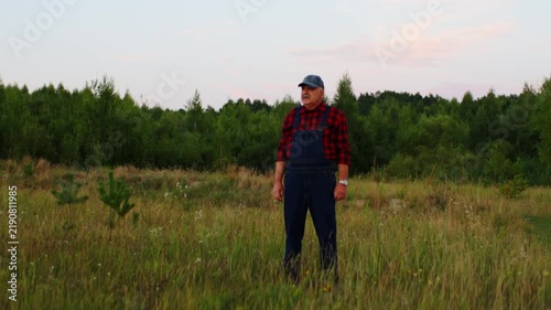 Elderly farmer in plaid shirt and overalls standing alone in tall grass field, contemplating nature at sunset near lush green forest horizon and open sky