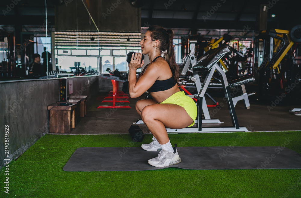Fototapeta premium Side view of determined Hispanic woman doing deep goblet squat with heavy dumbbell, athletic female performing lower body strength exercise in gym, leg day workout, copy space.