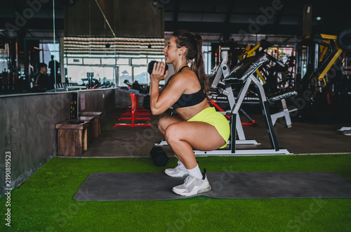 Fotografie Side view of determined Hispanic woman doing deep goblet squat with heavy dumbbell, athletic female performing lower body strength exercise in gym, leg day workout, copy space