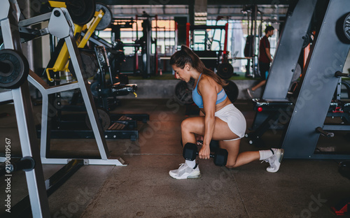 Obraz na płótnie Side view of athletic Hispanic woman performing dumbbell lunges in gym, fit female athlete training legs and glutes, functional strength exercise, body shaping workout, copy space