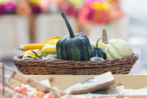 A wicker basket displays an assortment of colorful ornamental gourds and squashes in varied shapes, striped and curved, at an autumn farmers market stall with blurred flowers behind.
