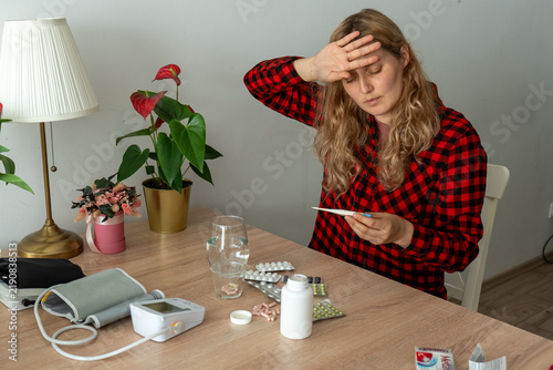 Unwell woman touching forehead and looking at thermometer beside medicines and blood pressure monitor. Concept of high fever, viral infection, home diagnosis and seasonal sickness.