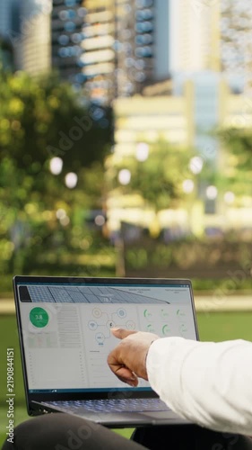 Portrait format: Businessman checks solar energy metrics and system data on a laptop outdoors in a city park, using high-tech sustainability data for a photovoltaics project.
