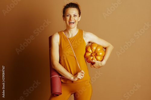 A cheerful woman with a broad smile looks at the camera, holding a yoga mat and an orange pumpkin, symbolizing autumn fitness and well-being.