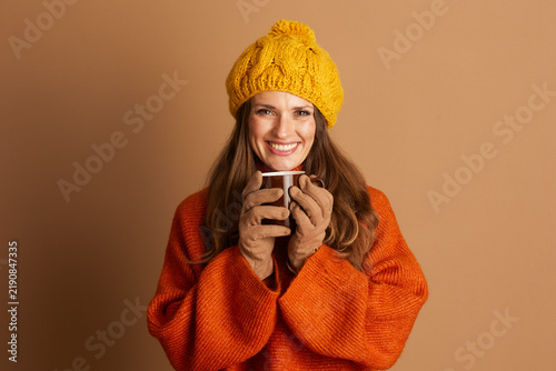 An adult woman with long hair, wearing yellow knitted pom-pom beanie and orange sweater, smiles directly at camera while holding brown mug. The cozy scene conveys cheerful autumn or winter atmosphere