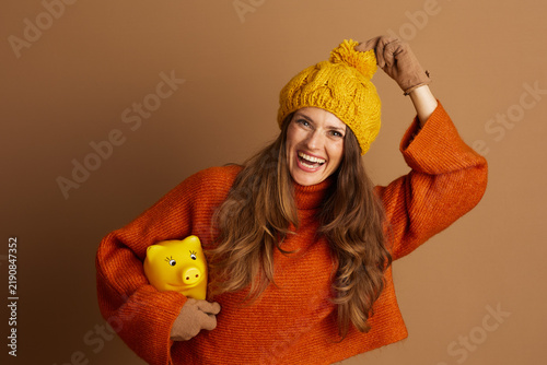 A joyful woman in orange sweater and yellow beanie laughs heartily while holding yellow piggy bank in her gloved hands. This image symbolizes financial planning and saving during cozy autumn season