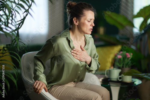 Принти на полотні A woman in a green satin blouse clutches her chest with an expression of distress, illustrating symptoms of anxiety, heart pain, or a panic attack at home