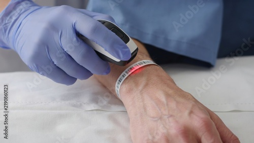 Barcode scanner in a gloved hand reading a patient wristband on an arm inside a clinic