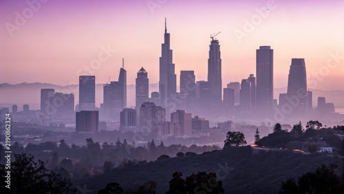 Long exposure of cityscape at dawn bathed in soft purple light with misty hills and tall skyscrapers creating calm and serene atmosphere