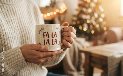 Cozy young woman holding a festive Christmas mug with Fa La La La text sitting in a warmly decorated living room during winter holidays