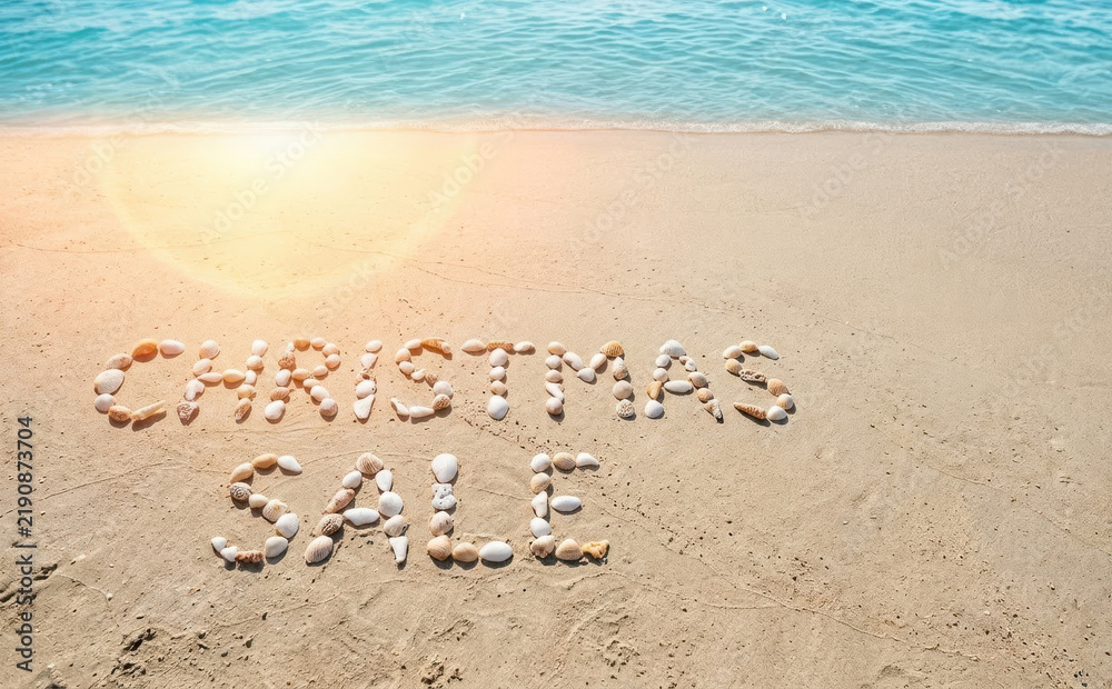 Fototapeta premium Christmas sale written with seashells on a tropical sandy beach with ocean waves for a tropical holiday shopping concept background