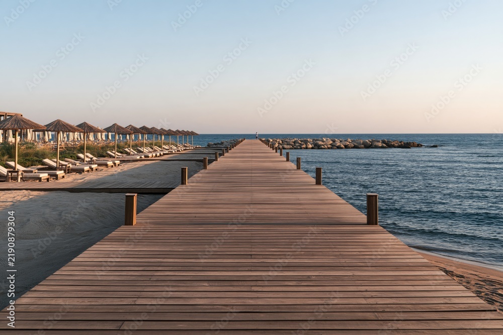 Obraz premium Wooden pier extending into the calm blue sea next to a sandy beach.
