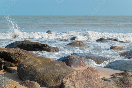 Waves crash against large dark rocks on coastal beach under clear blue sky