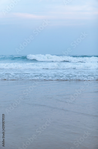 Gentle sea waves lap against wet sandy beach under pale blue sky