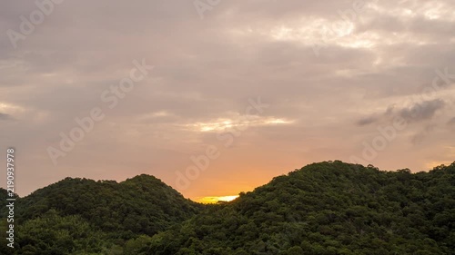 Time lapse view of a beautiful golden sunrise behind a lush green mountain range. Vibrant cloudy sky with orange and yellow morning sunlight over the forest landscape.